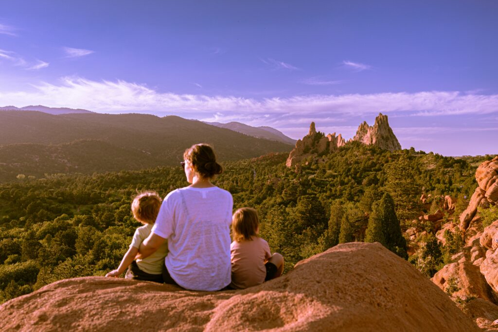 Family (mother and two children) sitting on a rock in Colorado's Garden of the Gods, looking towards scenic mountains and rock formations.