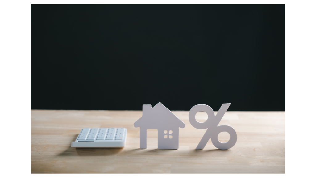 A white calculator, a house icon, and a percentage symbol on a wooden table against a dark background, illustrating mortgage rate calculations.